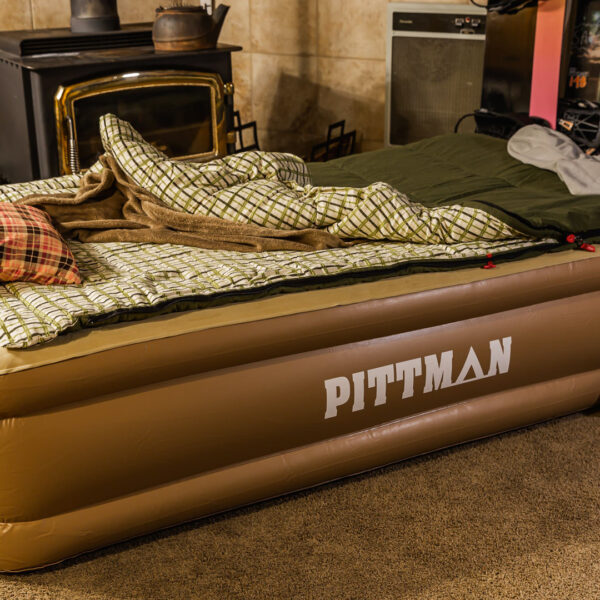 Lifestyle photo of inflated Pittman twin home mattress in room with fireplace, green and green and white plaid sleeping bag on top
